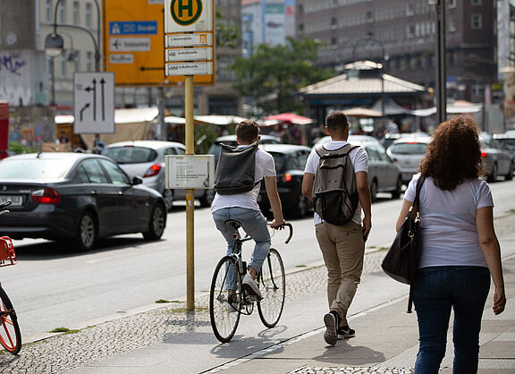 Eine Straßenszene: Ein Fahrradfahrer und zwei Fußgänger teilen sich einen gepflasterten Gehweg. Im Hintergrund parken Autos am Straßenrand.