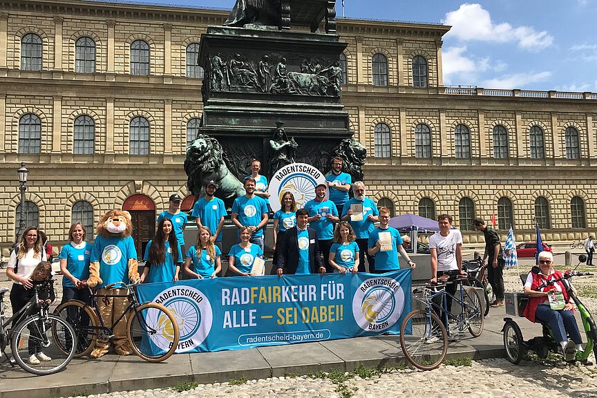 Auftakt zum Radentscheid Bayern Etwa 15 Personen in blauen Aktions-T-Shirts präsentieren auf einem Platz in München das Transparent des Radentscheids Bayern.